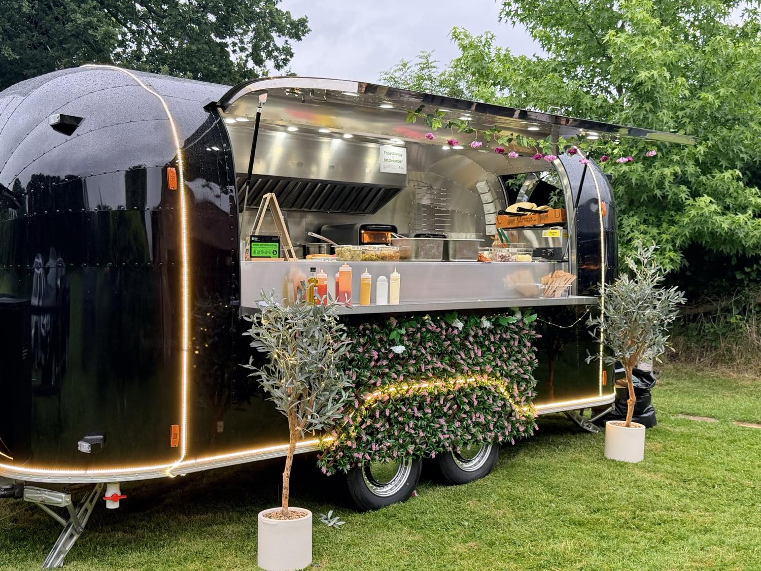 The ADevents 5-metre black Airstream kitchen with its serving hatch open, dressed with olive trees and flowers.