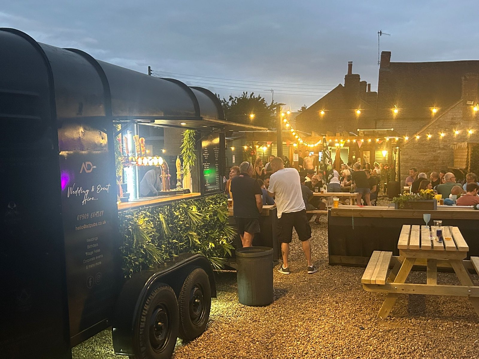 The ADevents horsebox bar serving a busy garden event under festoon lights at dusk.