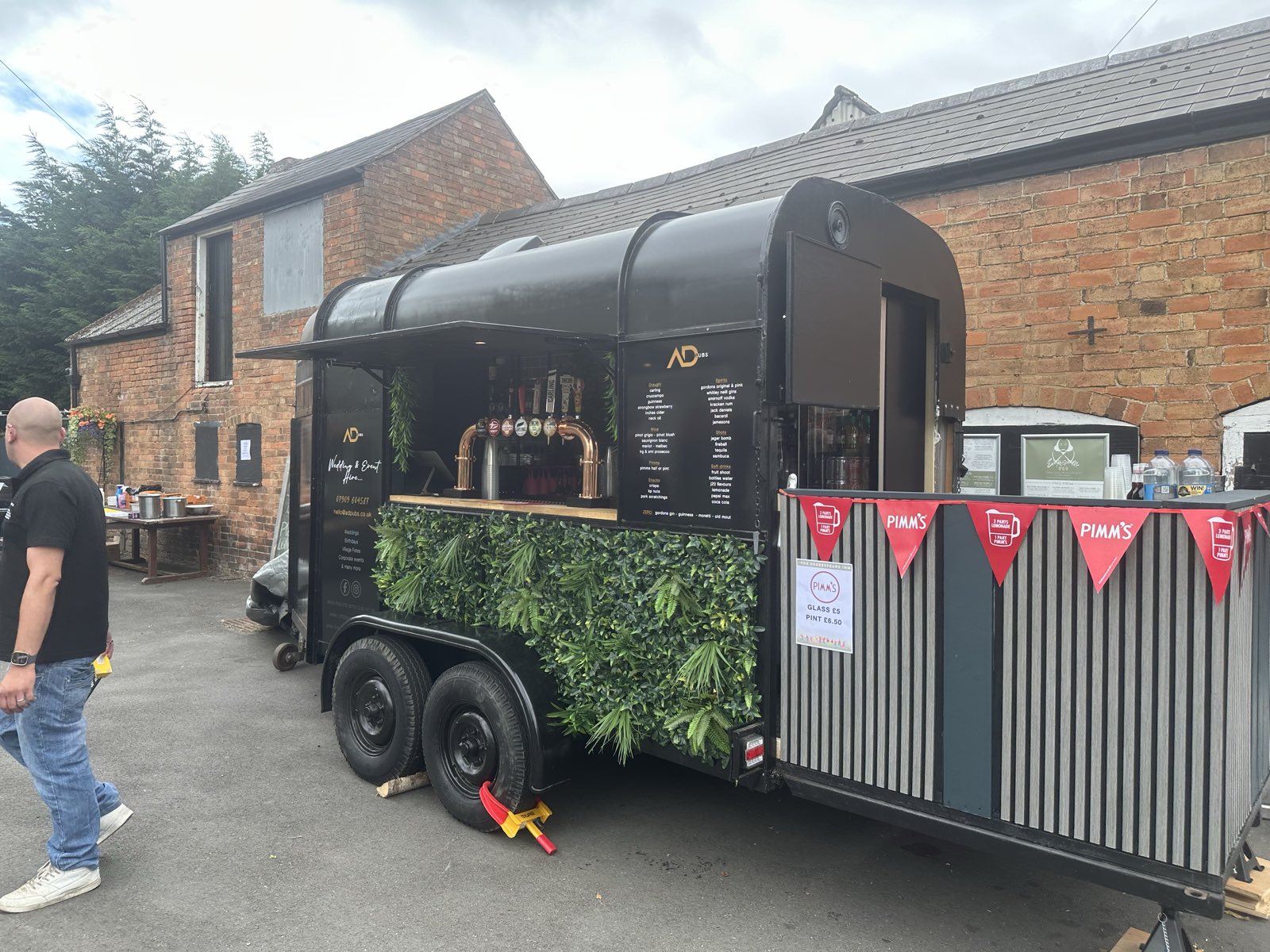 The black horsebox bar set up at an event beside a brick barn, bunting overhead.