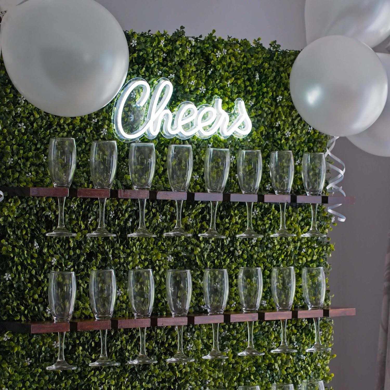 A prosecco glass wall with a neon Cheers sign and balloons, styled for a celebration.